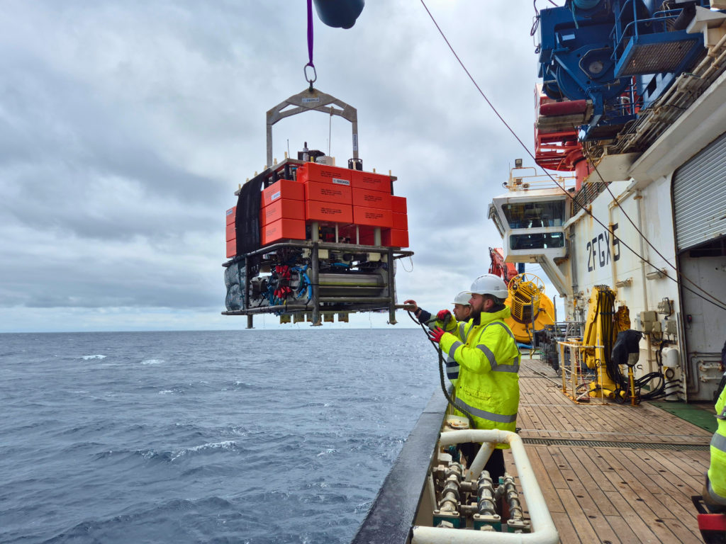 A large orange scientific instrument hangs from a crane beside a research vessel as crew members prepare for ocean deployment under cloudy skies