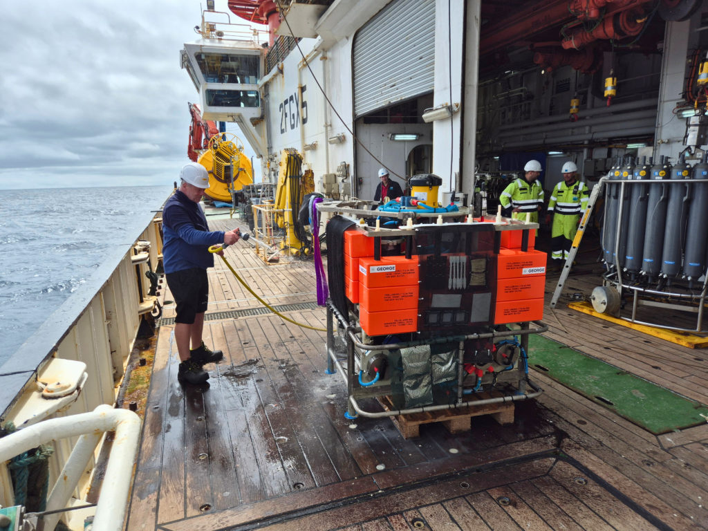 A crew member rinses a large orange ocean instrument with water on the deck of a research ship while other crew members stand nearby.