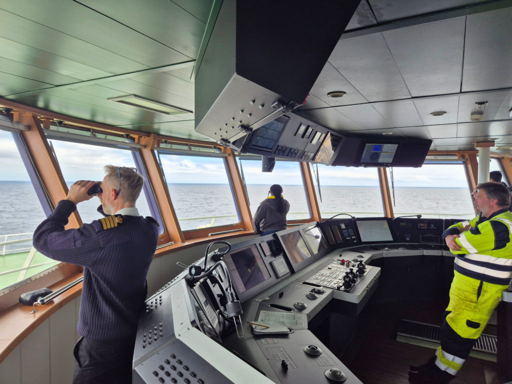 Crew members on the bridge of a research ship watch the ocean, with one person using binoculars and navigation consoles visible in the foreground